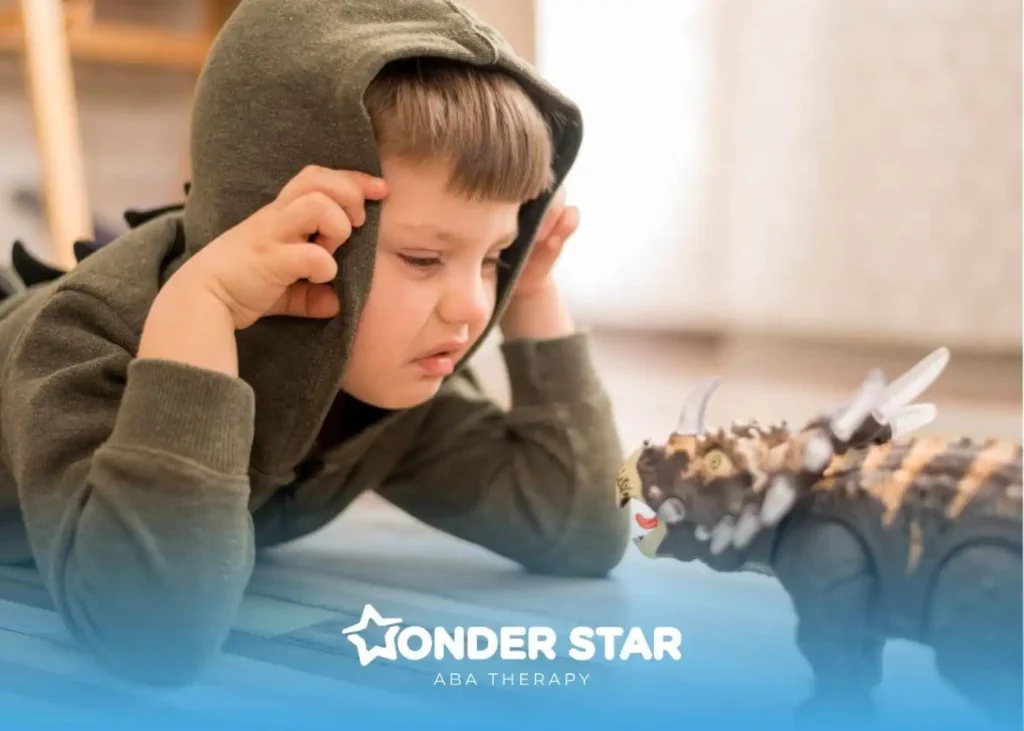An autistic boy in a hoodie holding ears while looking at a toy dinosaur, showing sensory sensitivity and focused attention.
