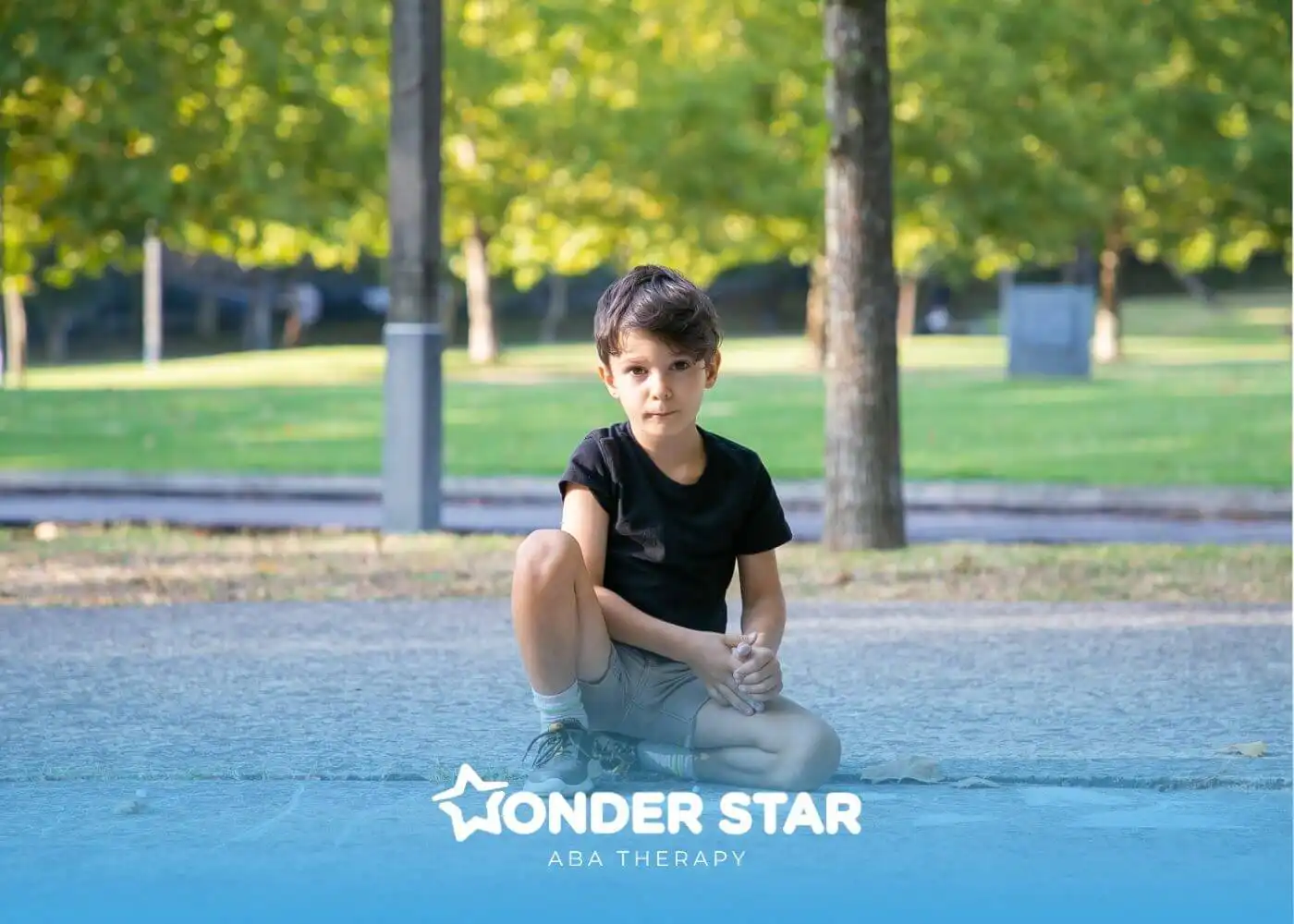 Young autistic boy sitting on pavement in a park, looking quietly at the camera with trees and grass behind in soft daylight.