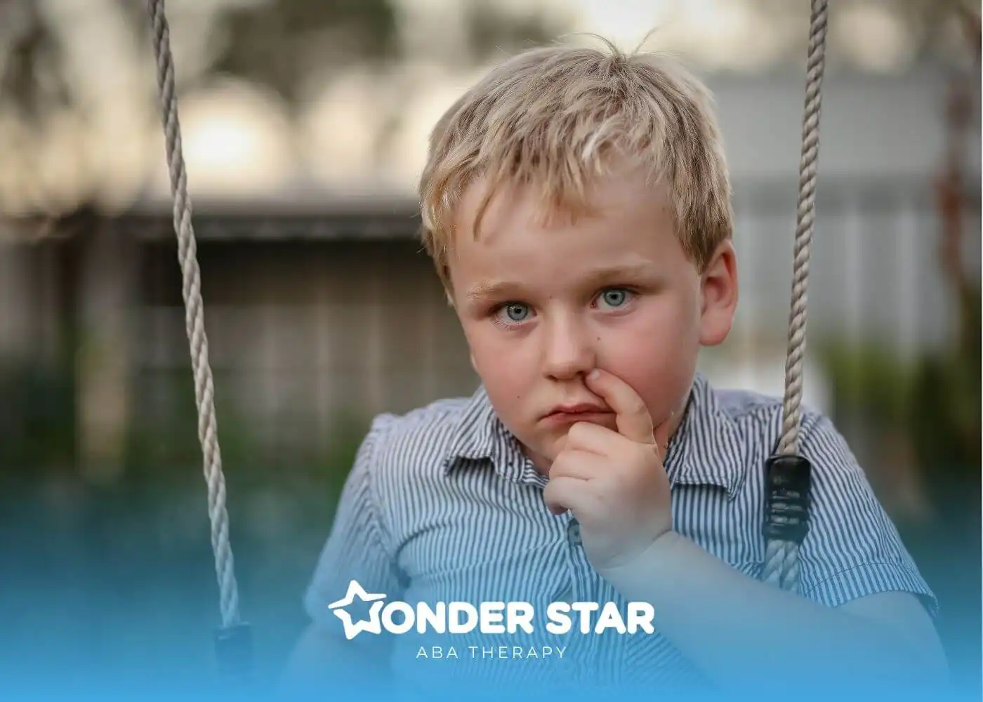 An autistic boy sitting on a swing with finger near nose, looking thoughtful and curious while playing in a backyard setting.