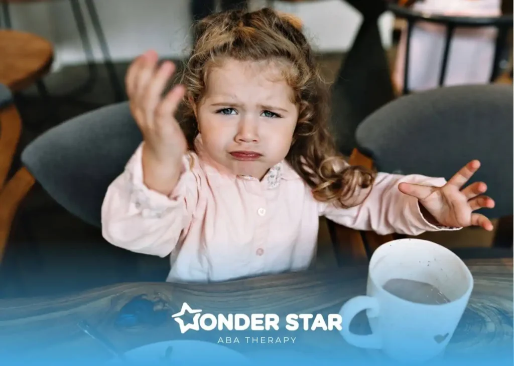 An autistic girl at a table is expressing frustration with hands raised, showing emotional regulation challenges.