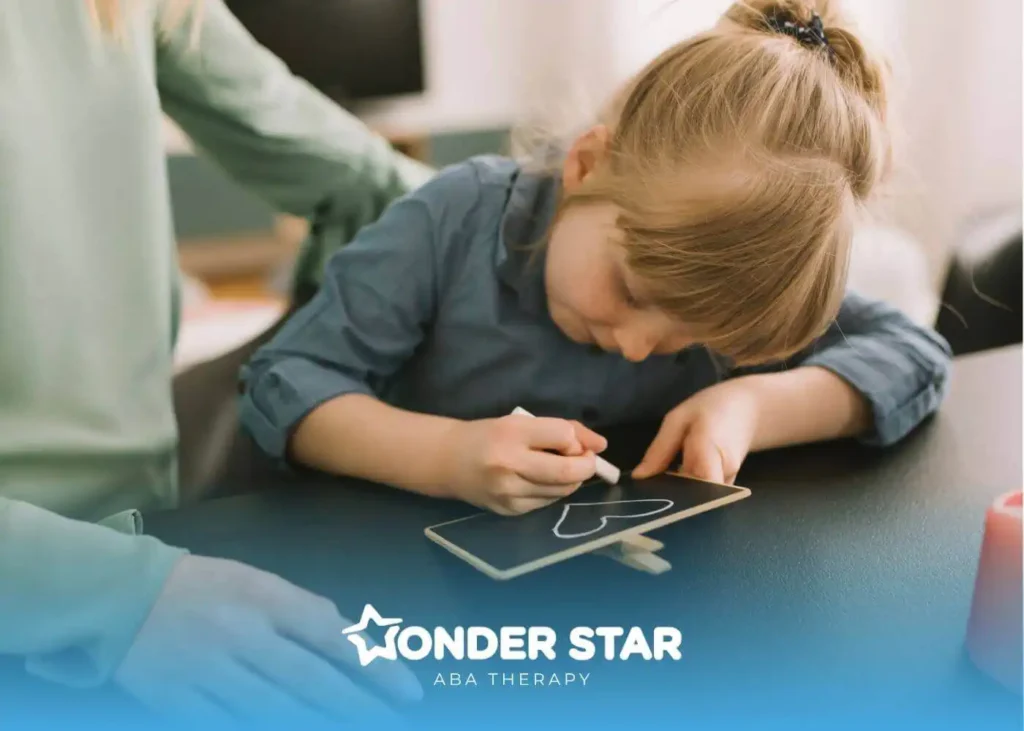 Young autistic girl drawing a heart with chalk on a small board while an adult supports her at a table.