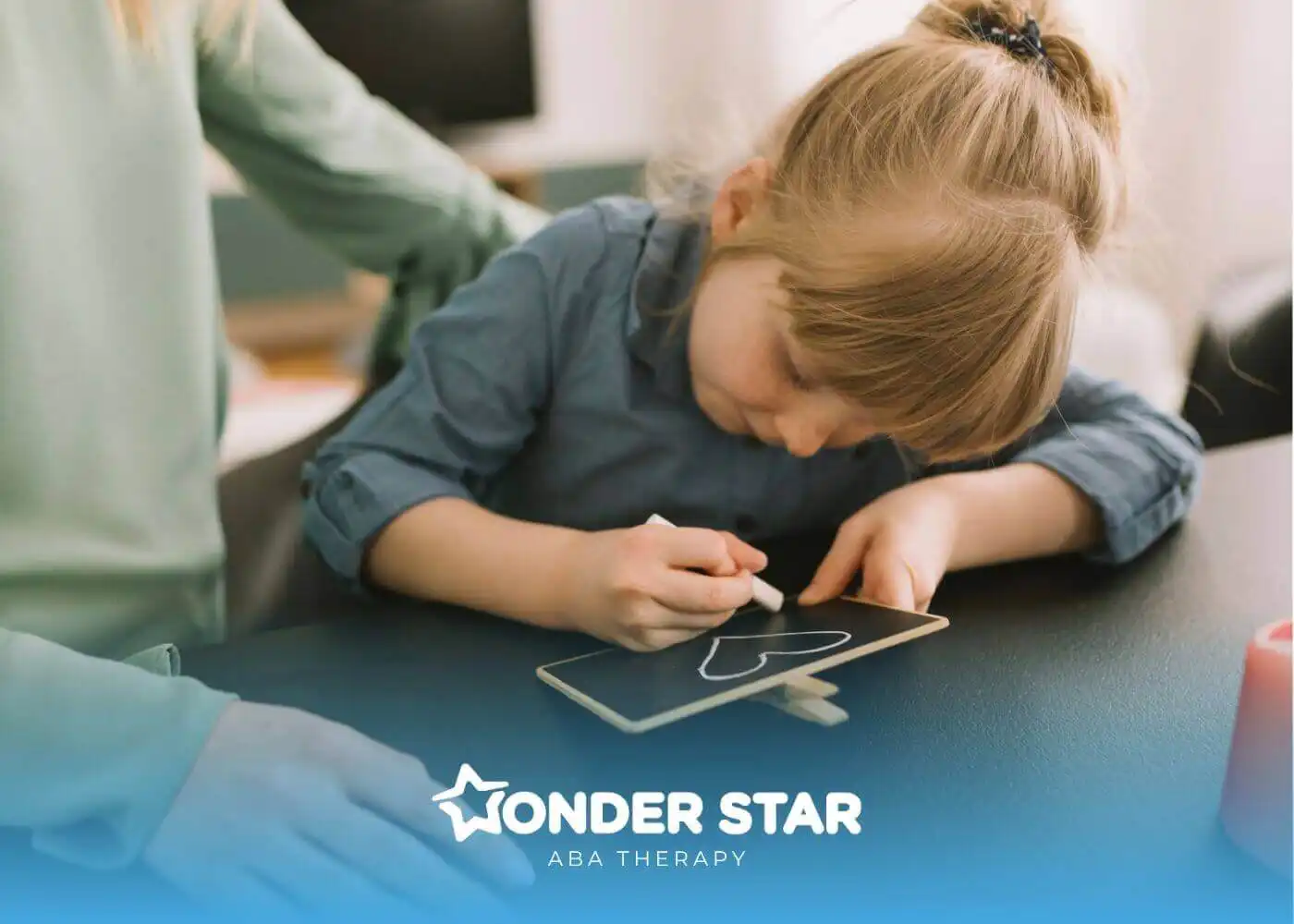 Young autistic girl drawing a heart with chalk on a small board while an adult supports her at a table.