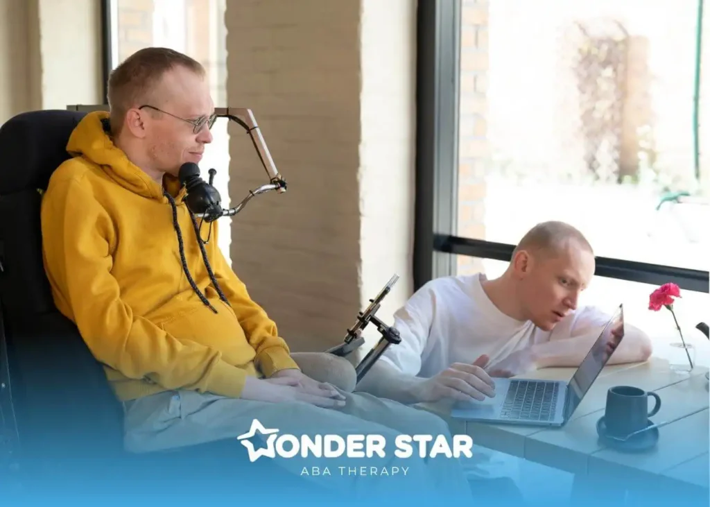 An autistic an in wheelchair using adaptive device at desk while another man works on laptop beside him.
