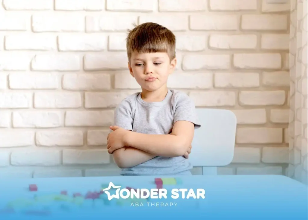 Autistic boy sitting with arms crossed at table with blocks, showing emotional response during structured activity session.