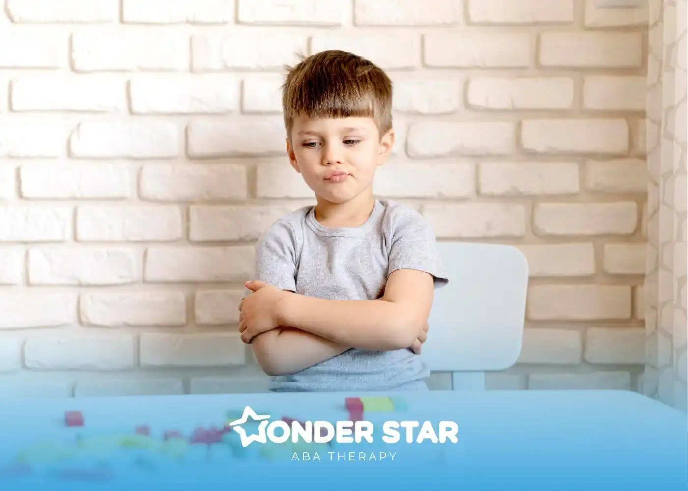 Autistic boy sitting with arms crossed at table with blocks, showing emotional response during structured activity session.