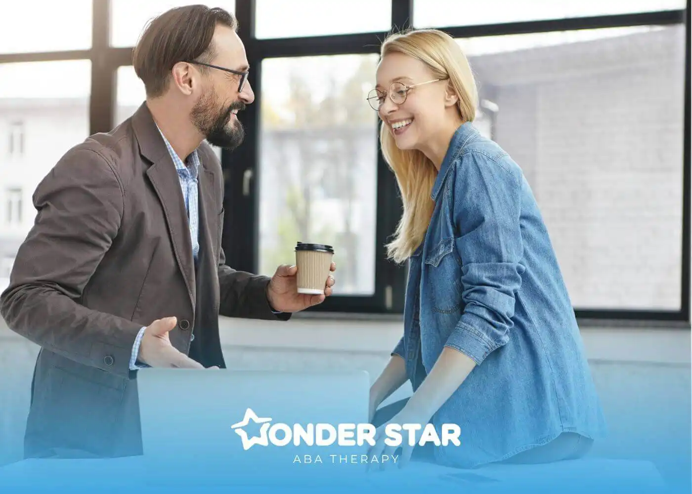 Two ABA therapist smiling and talking in an office, one holding a coffee cup beside a laptop.