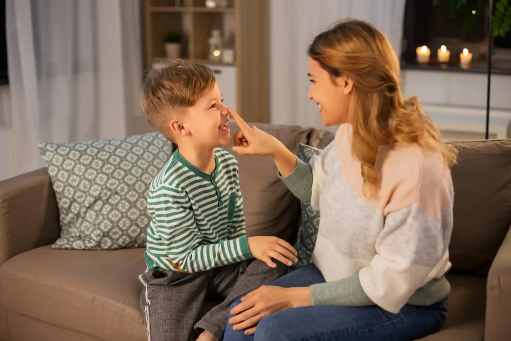 Smiling therapist gently touches nose of the autistic boy while they play together on a cozy couch at home.