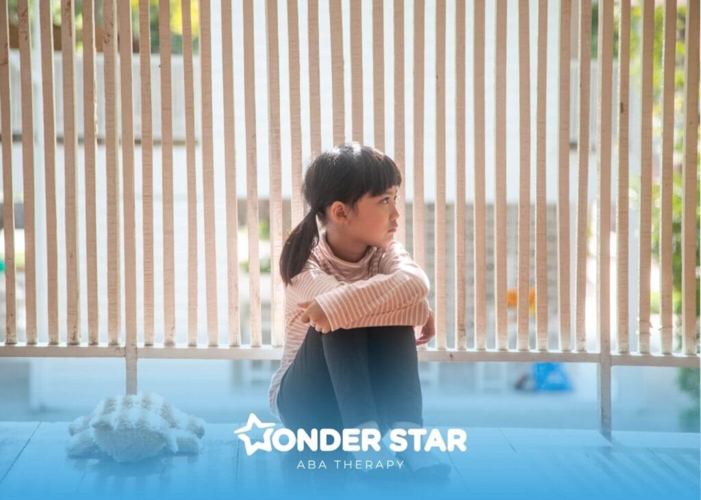Autistic girl sitting alone on a wooden floor near a railing looking outside with a stuffed toy beside her.